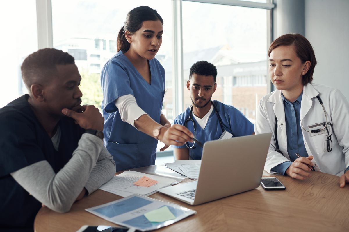 A group of early-career healthcare professionals, including two in scrubs and one in a white coat, are gathered around a table, focused on a laptop screen. One clinician is gesturing towards the laptop, seemingly explaining something to the group, while papers and a phone are spread out on the table. The group appears engaged in a collaborative discussion or training session in a well-lit medical office.